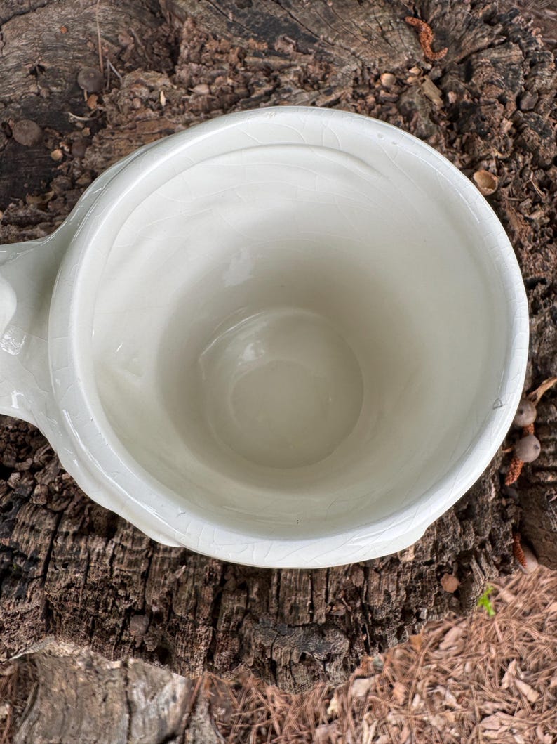 May include: A white ceramic planter with a rounded bowl shape and a small handle. The planter has a crackled glaze finish and is set against a natural wood background. The planter is empty.
