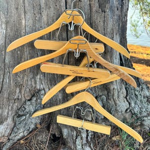 May include: A stack of wooden hangers with metal clips. The hangers are all natural wood and have a simple design. The hangers are stacked on top of each other and are hanging from a tree trunk.