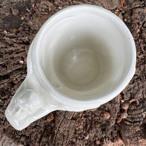 May include: Cream-colored ceramic mug with a sculpted face on the handle. The mug has a wide opening and a slightly textured surface. The mug is sitting on a weathered, brown tree stump.