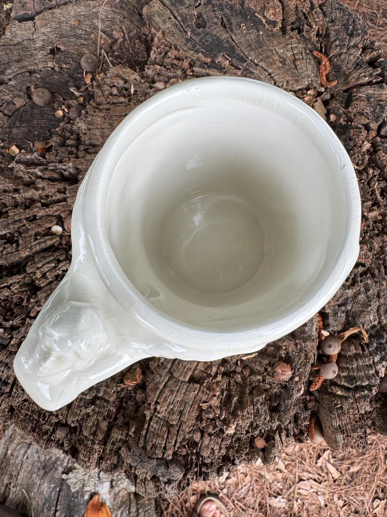 May include: Cream-colored ceramic mug with a sculpted face on the handle. The mug has a wide opening and a slightly textured surface. The mug is sitting on a weathered, brown tree stump.
