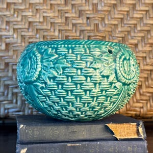 May include: A teal ceramic basket-shaped planter with a woven texture and floral accents. The planter sits on two antique books with dark blue covers. The background features a woven pattern.