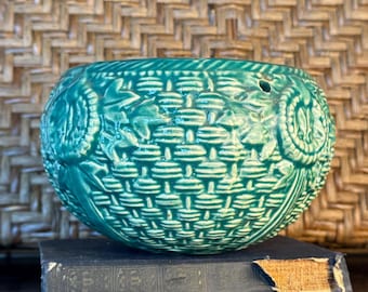 Pode incluir: Um vaso de cerâmica turquesa em forma de cesta, com textura tecida e detalhes florais. O vaso está sobre dois livros antigos com capas azul-escuras. O fundo apresenta um padrão tecido.
