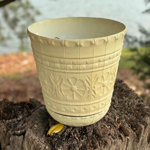 May include: A light yellow, decorative flower pot with embossed floral and geometric patterns. The pot has a wide opening and a slightly textured surface. The pot is sitting on a tree stump.
