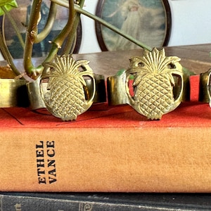 May include: Four brass pineapple napkin rings rest on a vintage book with the title "Reprisal" by Ethel Vance. The rings are gold-colored and feature a detailed pineapple design. The book is orange and sits atop a stack of other books.