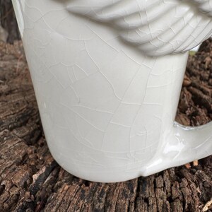 May include: A white ceramic mug with a wing-shaped design on the side. The mug has a crackle glaze finish, giving it a vintage appearance. The handle is curved and integrated into the mug's design.
