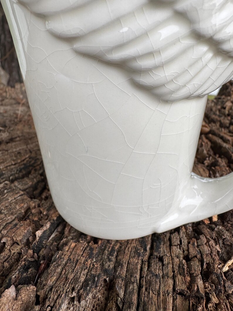 May include: A white ceramic mug with a wing-shaped design on the side. The mug has a crackle glaze finish, giving it a vintage appearance. The handle is curved and integrated into the mug's design.