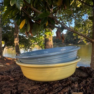 Puede incluir: Dos cuencos de cerámica, uno azul claro y otro amarillo, apilados sobre un tocón de árbol. Los cuencos tienen un diseño acanalado y están colocados sobre un fondo de lago y árboles, creando una estética rústica y natural.