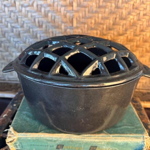 May include: A black ceramic flower frog bowl featuring a removable domed lid with a lattice grid pattern. The bowl has a smooth, glossy finish and small side handles, designed for arranging flowers with individual stems placed through the openings.