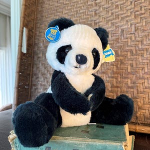 May include: A plush panda bear with black and white fur, featuring a round blue tag on the left ear and a yellow tag on the right ear, both displaying the Dakin logo. The panda is sitting upright with black limbs and a white face and torso.