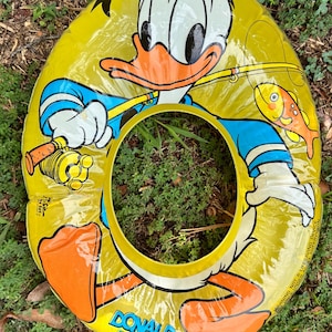 May include: A yellow inflatable swim ring featuring a graphic of Donald Duck holding a fishing rod with an orange fish nearby. The character wears a blue hat and shirt. The text DONALD DUCK is printed in blue at the bottom of the circular float.
