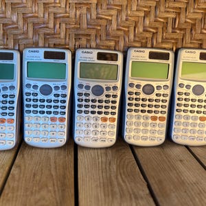 May include: Five Casio FX-115ES PLUS scientific calculators arranged in a row. Each calculator has a silver body, a green display screen, and a black and white button layout. The calculators are on a wooden surface.