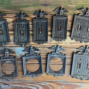 Puede incluir: Colección de cubiertas de interruptores de luz antiguas de color bronce, con un emblema de águila en la parte superior. Las cubiertas son rectangulares con varias configuraciones para interruptores simples y dobles, y algunas con aberturas circulares.