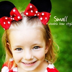 May include: A young girl smiles, wearing black mouse ears with red bows featuring white polka dots. The text "Small double bow style" is visible. The background is a blurred green.