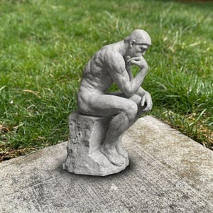 May include: A grey stone-colored sculpture of a seated figure in a contemplative pose, resting the chin on one hand. The figure is depicted with defined musculature, sitting on a textured, rock-like base with a weathered, matte finish.