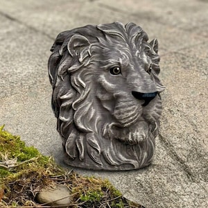 May include: A detailed, gray lion head sculpture with a textured finish. The lion's mane and facial features are intricately carved, and the eyes and nose are highlighted with darker tones. The sculpture is set on a stone surface.