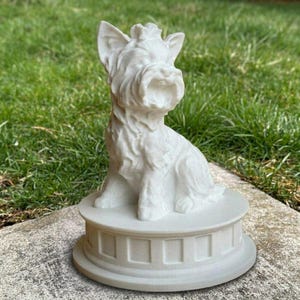 May include: A white matte figurine of a seated Yorkshire Terrier dog with pointed ears and textured fur detail. The dog is positioned on a circular, tiered pedestal base with a series of rectangular indentations around the side of the structure.