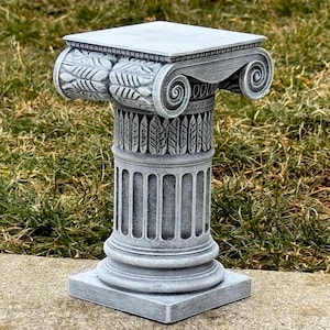 May include: A gray, decorative Ionic column with intricate carvings. The column features a square top, a detailed capital with scroll and leaf designs, and a fluted shaft. The base is also square. The column is made of a stone-like material.