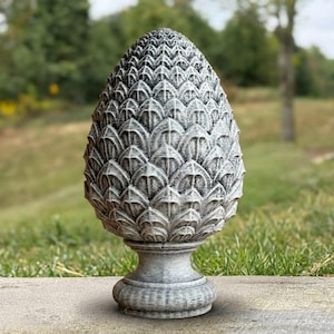 May include: A gray and white decorative finial shaped like a pinecone with a textured, layered scale pattern. The object features a rounded, pointed top and sits on a circular, tiered pedestal base with a weathered, stone-like finish throughout.