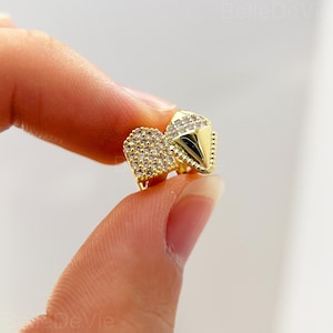 May include: A pair of gold-colored stud earrings. One earring is heart-shaped and covered in small, clear crystals. The other earring is a pyramid shape with a pointed tip and also features clear crystals.