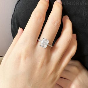 May include: A silver ring featuring a large, square-shaped diamond in the centre. The diamond is surrounded by smaller diamonds that form a halo around it. The ring is set on a thin band.