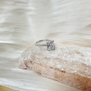 May include: A radiant, square-shaped diamond engagement ring with a pave setting on the band. The ring is set on a light brown rock.