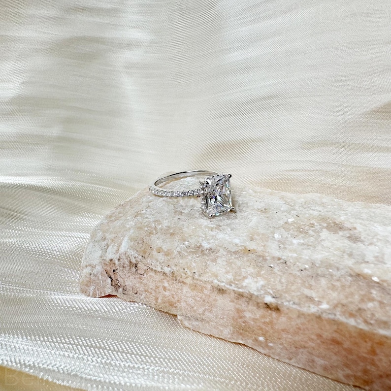 May include: A radiant, square-shaped diamond engagement ring with a pave setting on the band. The ring is set on a light brown rock.