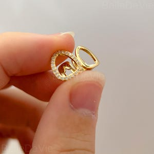 Pode incluir: Um brinco dourado com um design de argola dupla. Uma argola é incrustada com pequenos cristais transparentes, a outra tem uma forma lisa e aberta. O brinco é segurado em uma mão, mostrando seus detalhes.
