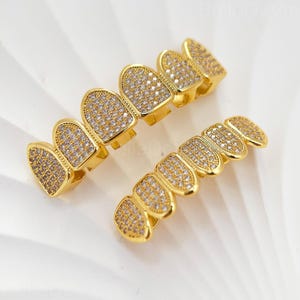 May include: Two sets of gold-coloured, jewelled teeth grillz. Each grillz set is encrusted with small, clear gemstones. The top set has six teeth, and the bottom set has six teeth. The grillz are displayed on a white surface.