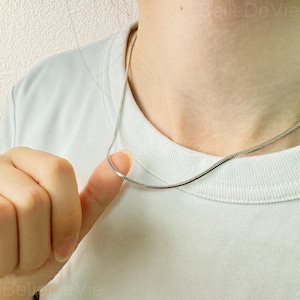 May include: A silver snake chain necklace. The chain is thin and delicate, and it is worn by a person with a white shirt.