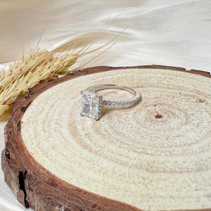 May include: A silver ring with a large, square-shaped diamond set in a halo of smaller diamonds. The ring is on a wooden surface.