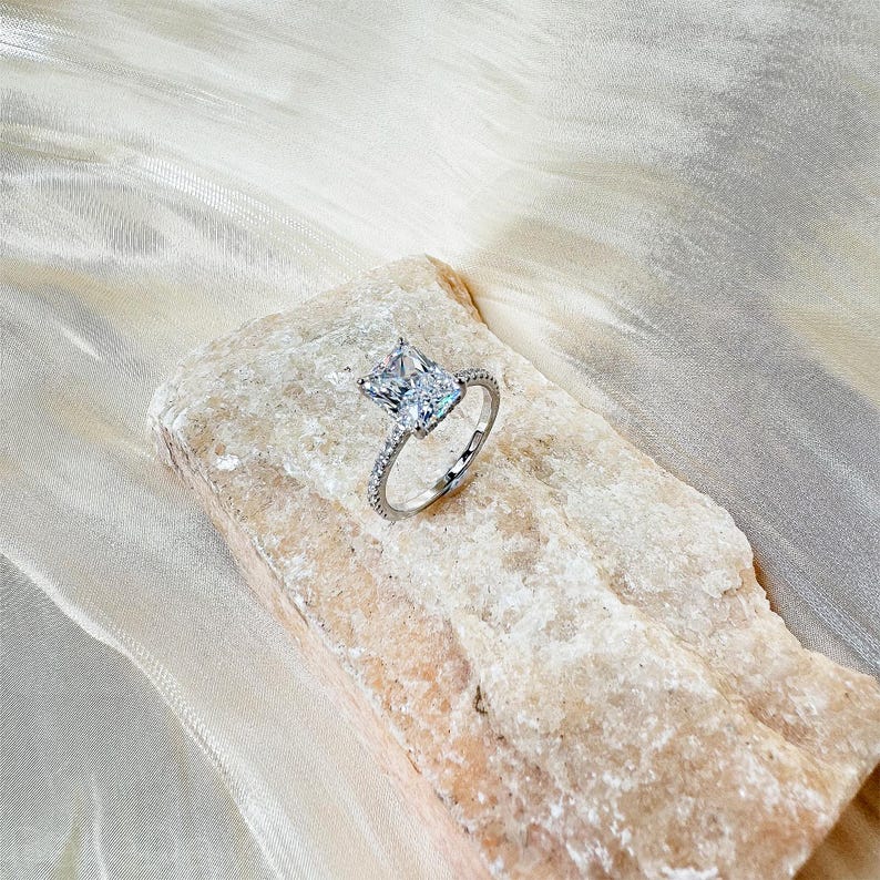 May include: A silver ring with a large, clear, square-shaped diamond. The ring is set on a rock-like surface.