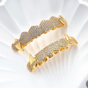 May include: Two gold-colored, custom-fit teeth grillz. Each grill is encrusted with small, sparkling, clear stones. The grillz have a unique design with pointed edges and are displayed on a white surface.