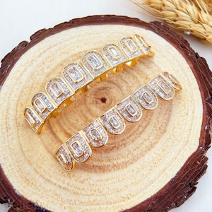 May include: Two gold-colored grillz, one for the top teeth and one for the bottom, are displayed on a wooden surface. Each grill is encrusted with rectangular, clear gemstones. The grillz have a curved, scalloped design.