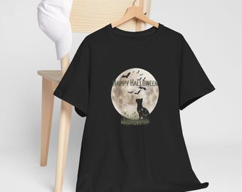 May include: Black t-shirt featuring a Halloween design. The graphic includes a full moon, bats, and a cat silhouette. The words "Happy Halloween" are printed above the moon. A white baseball cap rests on a wooden chair.