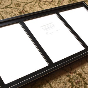 May include: A black triple picture frame with white matting. The frame has a thin gold accent line. The frame is on a patterned rug. The frame is empty and ready for photos.