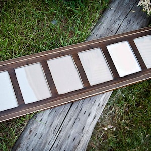 May include: A rectangular, multi-photo frame with six openings. The frame is made of dark brown wood with a lighter trim. The frame is resting on a weathered wooden surface with green grass in the background.