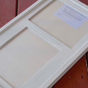 May include: White wooden picture frame with three openings. The frame has a distressed white finish and a slightly raised edge around each opening. The frame is sitting on a red wooden surface.