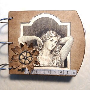 May include: A handmade scrapbook with a vintage aesthetic. The cover is brown with a black and white illustration of a woman. A decorative flower made of brown paper and a gear is attached to the cover. The book is bound with silver rings.