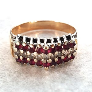 May include: A gold ring with three rows of gemstones. The top and bottom rows feature small, round, deep red stones, while the middle row has smaller, clear stones. The ring has a classic, elegant design.