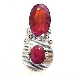 May include: A silver pendant with two oval, deep red gemstones. The larger gem is at the top, the smaller one below, set in a textured silver base. Decorative silver accents complete the design.