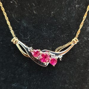 May include: A gold-coloured necklace with a silver and gold pendant. The pendant features three heart-shaped, bright pink gemstones. The necklace has a delicate chain and is set against a dark background.