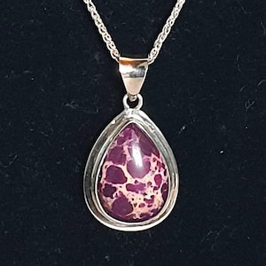 May include: A silver necklace featuring a teardrop-shaped pendant. The pendant has a purple and cream-colored stone set in a silver bezel. The chain is a delicate silver cable chain, perfect for everyday wear.