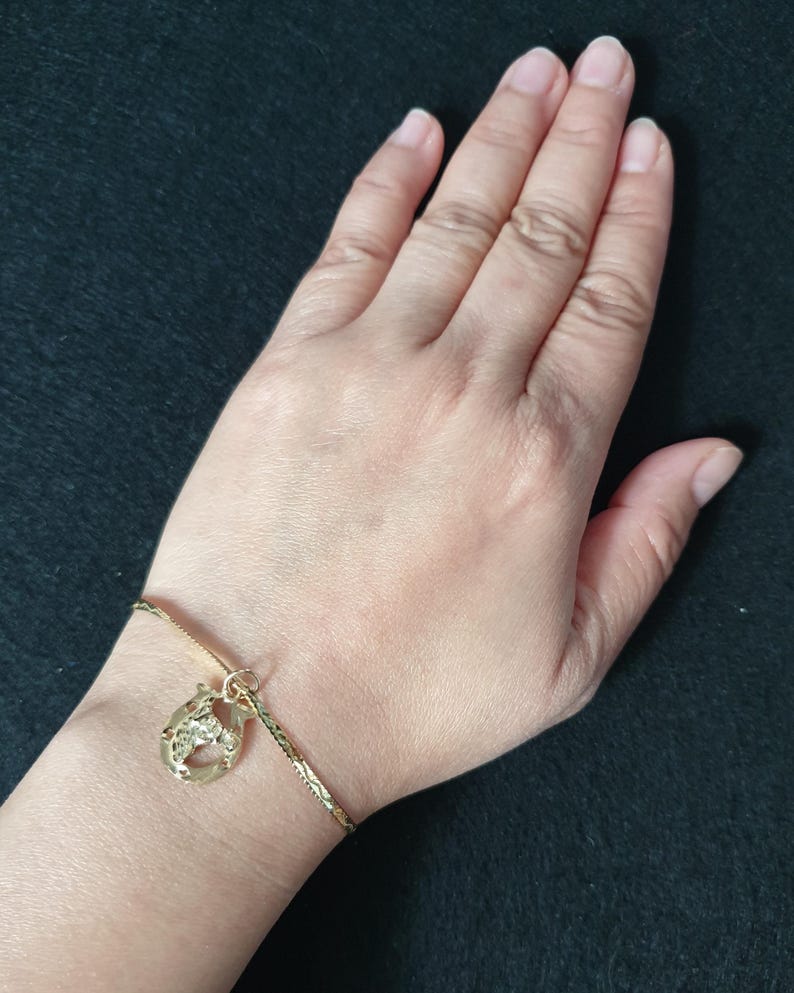 May include: A gold-toned bracelet featuring a textured chain and a single horseshoe-shaped charm. The charm has a detailed, embossed surface and is attached to the bracelet with a small circular jump ring, creating a simple and classic jewellery piece.