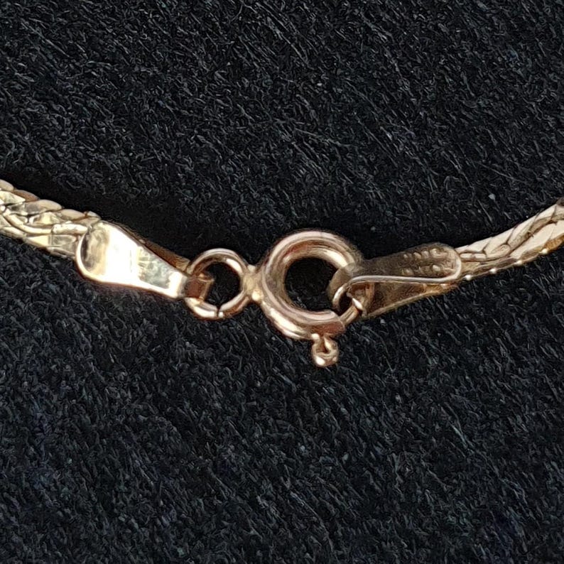 May include: A gold-toned chain necklace featuring a circular spring ring clasp mechanism. The metal links exhibit a woven, textured pattern that connects to the polished, smooth metal components of the fastener on both ends of the chain.