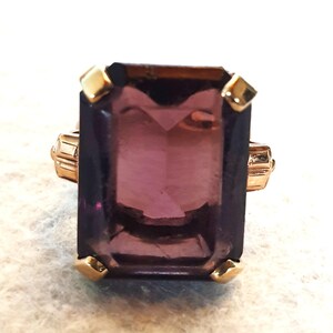 May include: A gold ring with a large, rectangular, purple gemstone. The corners of the gemstone are secured with small, gold-coloured prongs. The ring's band has a detailed, geometric design.