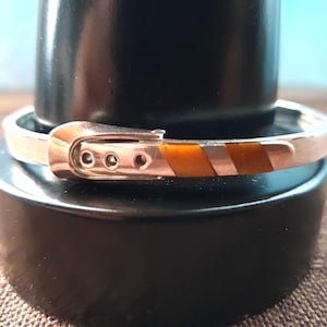 May include: Silver bracelet with a buckle design. The bracelet has a silver band with a buckle clasp and brown and white stripes. The bracelet is displayed on a black stand.
