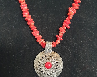 Puede incluir: Un collar de coral rojo con un colgante de plata que presenta un diseño circular y una piedra preciosa roja en el centro.