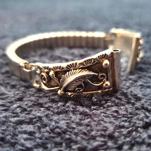 May include: Silver watch strap with an intricate design. The strap has a detailed feather and scrollwork pattern on the decorative central section. The band is made of a flexible, expandable material.