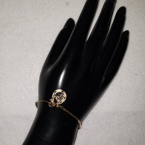 May include: A gold-toned chain bracelet featuring a small horseshoe-shaped charm. The charm has a textured surface and is attached to a delicate, thin metal chain that wraps around the wrist. The metal has a polished, reflective finish.