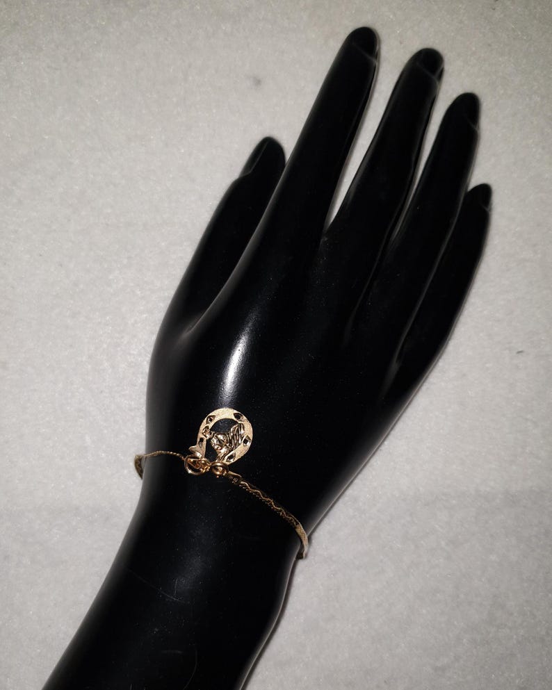 May include: A gold-toned chain bracelet featuring a small horseshoe-shaped charm. The charm has a textured surface and is attached to a delicate, thin metal chain that wraps around the wrist. The metal has a polished, reflective finish.